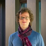 Headshot of a white man with short curly brown hair and glasses, wearing a blue sweater and maroon scarf.