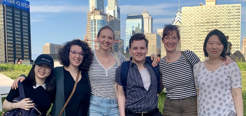 Six happy linguists from the lab stand together in front of the Philadelphia skyline. There are two Asian women, three white women, and one white man, arm in arm.