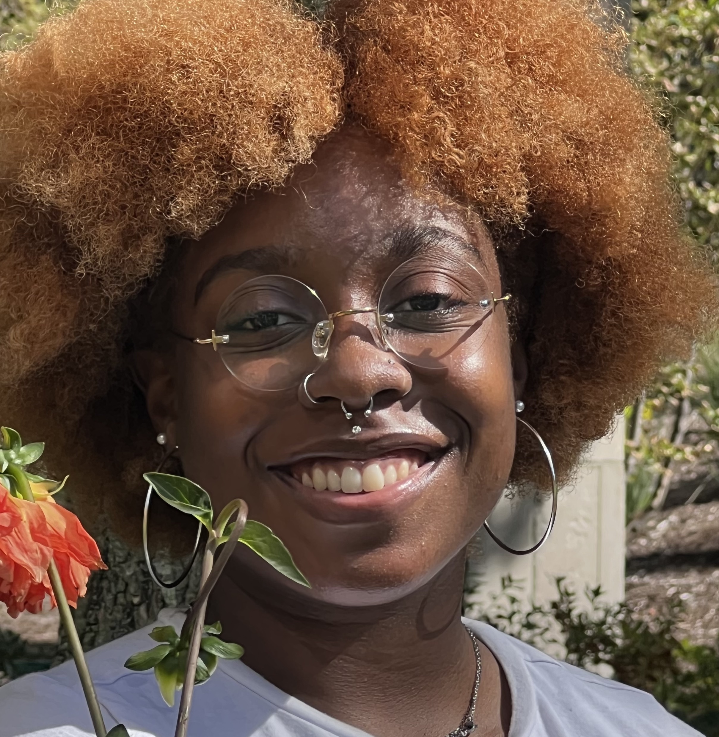 Headshot of a Black non-binary person with glasses and piercings standing behind a flower.